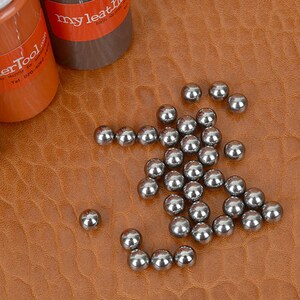 May include: A collection of small, shiny metal ball bearings scattered on a textured, brown leather surface. Three bottles of leather tool products are visible in the background, with labels that read "my leatherTool".