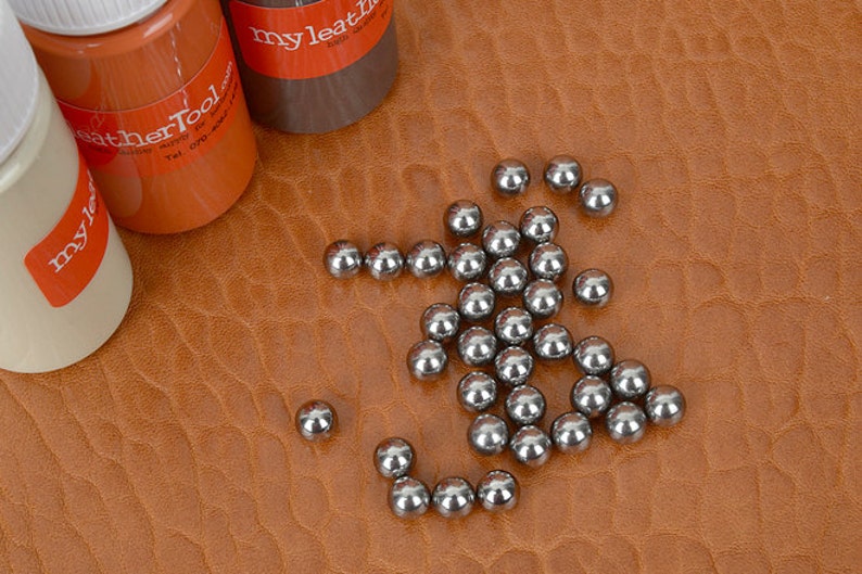 May include: A collection of small, shiny metal ball bearings scattered on a textured, brown leather surface. Three bottles of leather tool products are visible in the background, with labels that read "my leatherTool".