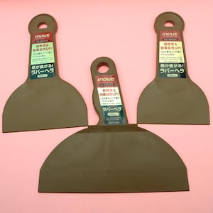 May include: Three brown rubber scrapers with rounded blades and a hole in the handle. Each scraper has a label with Japanese text and measurements of 100mm, 125mm, and 200mm. The background is a solid pink.