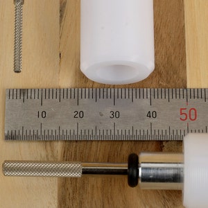 May include: An overhead shot shows a white plastic component, a metal rod with a textured handle, and a metal ruler with centimeter markings. The ruler measures up to 60 cm. The objects are on a light wood surface.