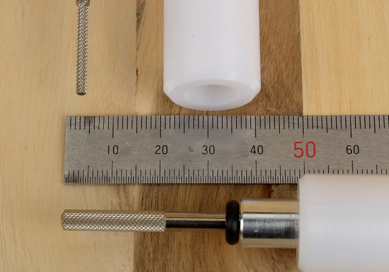 May include: An overhead shot shows a white plastic component, a metal rod with a textured handle, and a metal ruler with centimeter markings. The ruler measures up to 60 cm. The objects are on a light wood surface.