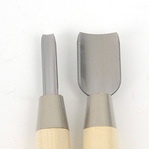 Op de afbeelding: Twee houtsnijgereedschappen met metalen bladen. Eén gereedschap heeft een recht blad, de andere een gebogen blad. Beide hebben lichtgekleurde houten handvatten en zilverkleurige metalen hulzen. De gereedschappen staan tegen een witte achtergrond.