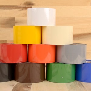 May include: A collection of brightly coloured tape rolls, arranged in a pyramid shape. The colours include white, yellow, red, orange, grey, black, brown, green, and blue. The background is light brown wood.
