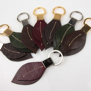 Pu&ograve; includere: Una collezione di portachiavi a forma di foglia in vari colori, tra cui marrone, verde e bordeaux. Ogni portachiavi presenta un anello metallico e una cucitura decorativa al centro, con dettagli dorati o argentati.