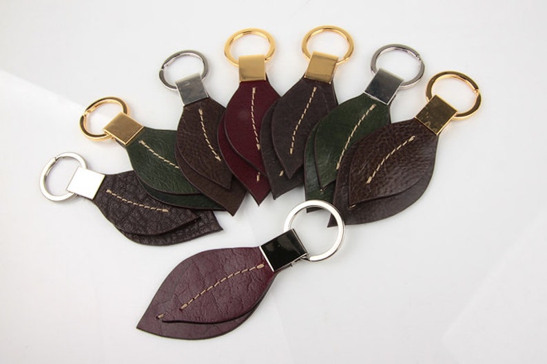 Pu&ograve; includere: Una collezione di portachiavi a forma di foglia in vari colori, tra cui marrone, verde e bordeaux. Ogni portachiavi presenta un anello metallico e una cucitura decorativa al centro, con dettagli dorati o argentati.
