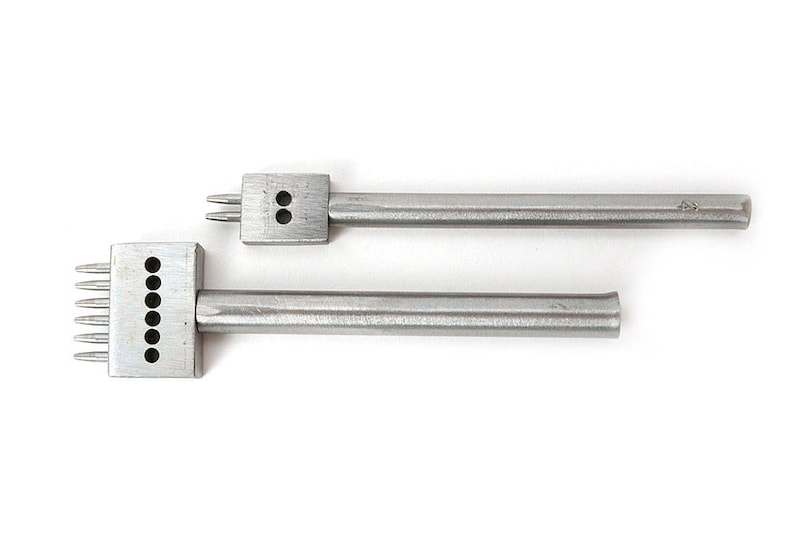 May include: Two silver leatherworking tools. One has two prongs and two holes, the other has six prongs and six holes. Both tools have a rectangular head and a long, cylindrical handle. They are designed for creating evenly spaced holes in leather.