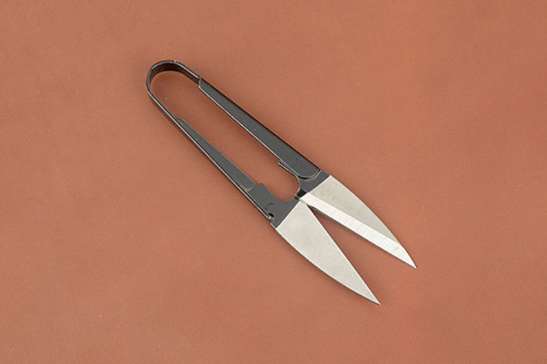 K&ouml;nnte beinhalten: Eine Schere mit schwarzen Griffen und silbernen Klingen, wahrscheinlich f&uuml;r Bastelarbeiten oder Gartenarbeit, liegt auf einer braunen Oberfl&auml;che. Die Klingen sind scharf und angewinkelt, und die Griffe bilden eine Schlaufe. Die Schere ist f&uuml;r pr&auml;zises Schneiden konzipiert.
