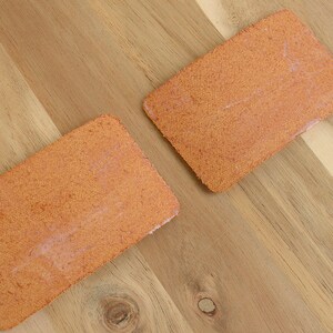 May include: Two rectangular, orange-colored, textured pads are displayed on a wooden surface. The pads have a slightly rough texture and appear to be made of a woven material. The wooden surface has a natural wood grain pattern.