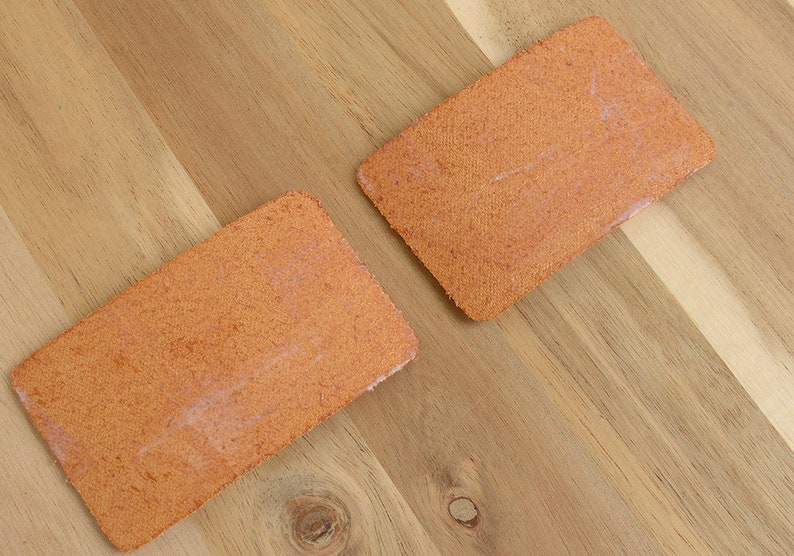 May include: Two rectangular, orange-colored, textured pads are displayed on a wooden surface. The pads have a slightly rough texture and appear to be made of a woven material. The wooden surface has a natural wood grain pattern.