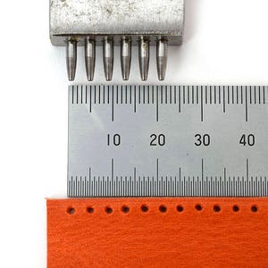 May include: A metal hole punch tool with multiple cylindrical punches is positioned above a ruler and a strip of orange material with punched holes. The ruler shows measurements in centimetres.