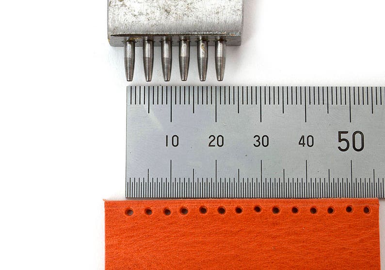 May include: A metal hole punch tool with multiple cylindrical punches is positioned above a ruler and a strip of orange material with punched holes. The ruler shows measurements in centimetres.