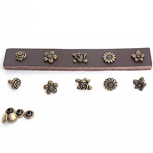 May include: A dark brown leather strip featuring five antique brass-toned metal flower-shaped rivets attached in a row. Below, five matching individual flower rivets are arranged horizontally, with four additional plain metal rivet backings.