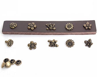 May include: A dark brown leather strip featuring five antique brass-toned metal flower-shaped rivets attached in a row. Below, five matching individual flower rivets are arranged horizontally, with four additional plain metal rivet backings.