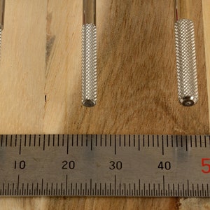 May include: Three metal tools with textured handles are displayed against a wooden background. A metal ruler with centimeter markings is placed below the tools. The tools have a silver finish and are of varying lengths.