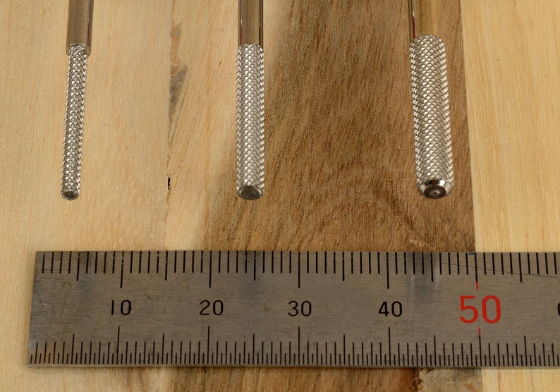 May include: Three metal tools with textured handles are displayed against a wooden background. A metal ruler with centimeter markings is placed below the tools. The tools have a silver finish and are of varying lengths.