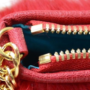 May include: Close-up of a red handbag with a gold chain strap and zipper. The bag has a textured leather exterior and a fluffy, fur-like texture. The interior is teal. The zipper is gold.