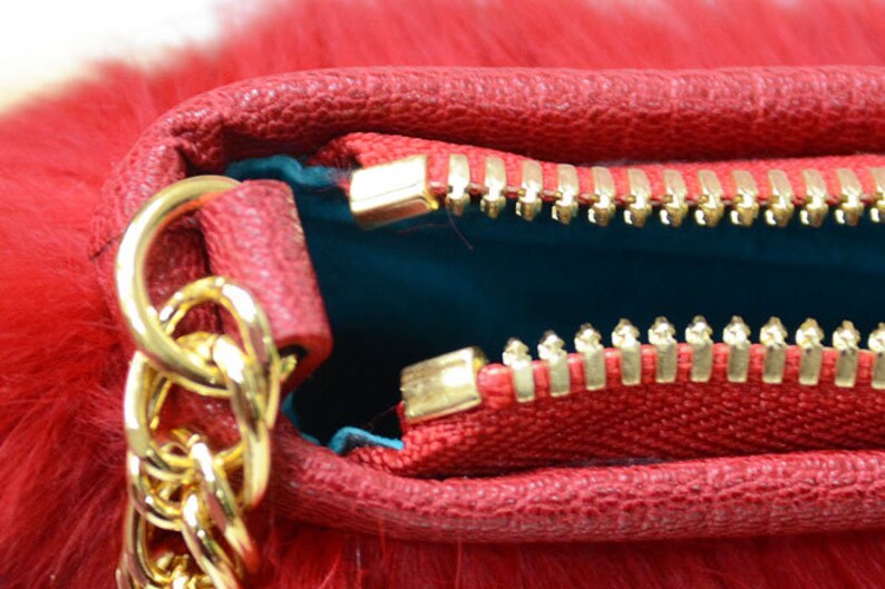 May include: Close-up of a red handbag with a gold chain strap and zipper. The bag has a textured leather exterior and a fluffy, fur-like texture. The interior is teal. The zipper is gold.