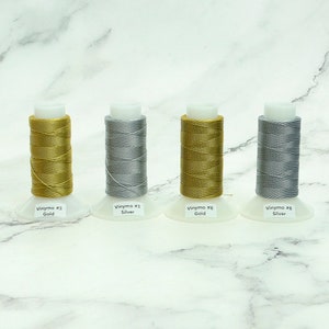 May include: Four spools of Vinymo thread in gold and silver colors. Each spool is labeled with its color and the Vinymo number. The spools are on white bases, and the background is a marble pattern.