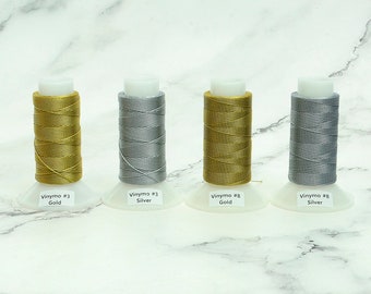 May include: Four spools of Vinymo thread in gold and silver colors. Each spool is labeled with its color and the Vinymo number. The spools are on white bases, and the background is a marble pattern.