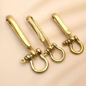 May include: Three brass keychains with a hook and a D-ring. The keychains are made of shiny, gold-colored metal. The hooks are curved and the D-rings are round. The keychains are arranged in a row on a light beige surface.
