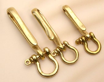 May include: Three brass keychains with a hook and a D-ring. The keychains are made of shiny, gold-colored metal. The hooks are curved and the D-rings are round. The keychains are arranged in a row on a light beige surface.