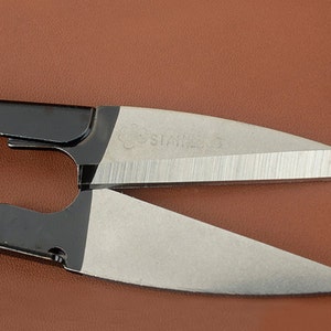 K&ouml;nnte beinhalten: Nahaufnahme einer Schere aus Edelstahl mit schwarzen Griffen. Die silbernen Klingen tragen die Gravur "STAINLESS". Die Schere befindet sich auf einem braunen Hintergrund.