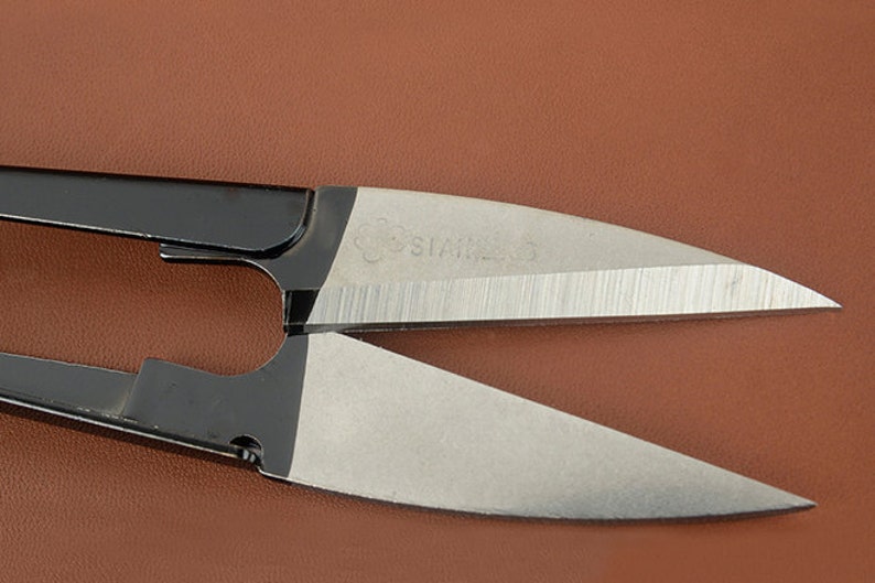 K&ouml;nnte beinhalten: Nahaufnahme einer Schere aus Edelstahl mit schwarzen Griffen. Die silbernen Klingen tragen die Gravur "STAINLESS". Die Schere befindet sich auf einem braunen Hintergrund.