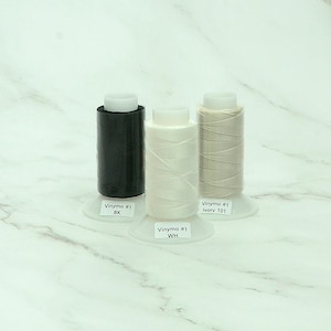 May include: Three spools of Vinymo #1 thread in black, white, and ivory. Each spool is labeled with its color and sits on a clear plastic base. The thread is tightly wound, and the spools are arranged against a white marble background.