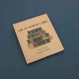 Könnte beinhalten: Ein beigefarbenes Heft mit dem Titel "OIL LEATHER CORD" und einer Illustration von Garnrollen in verschiedenen Farben. Der Text "천연 가죽끈 Genuine Leather" ist darunter gedruckt. Das Heft liegt auf einer blauen Oberfläche.