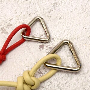 May include: Close-up of two leather keychains with silver-colored metal carabiner clips. One keychain is red, and the other is a pale yellow. The leather is knotted, and the clips are triangular.