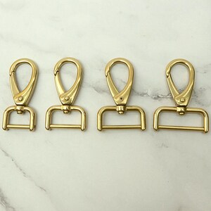 Pode incluir: Quatro fechos giratórios dourados com base em forma de D. Os fechos têm um acabamento liso e polido e são projetados para serem fixados a bolsas ou outros acessórios. Os fechos estão dispostos em uma fileira contra um fundo de mármore branco.