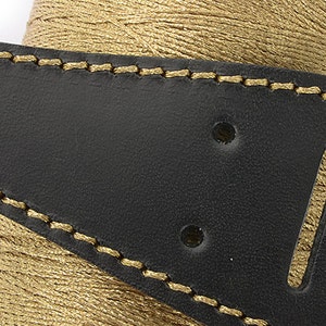 May include: Close-up of a black leather guitar strap with gold stitching and a gold thread spool in the background. The strap has two holes and a slot for adjustment. The stitching is detailed and the leather has a smooth texture.