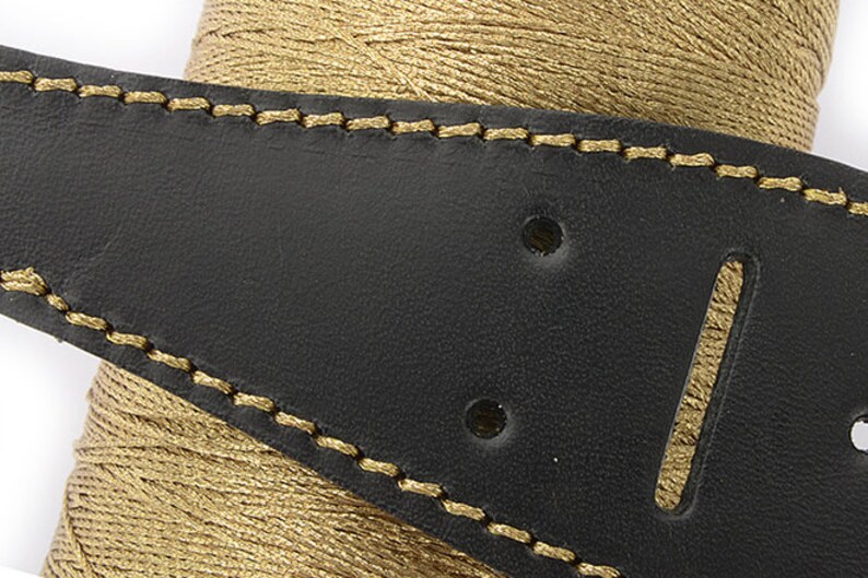 May include: Close-up of a black leather guitar strap with gold stitching and a gold thread spool in the background. The strap has two holes and a slot for adjustment. The stitching is detailed and the leather has a smooth texture.