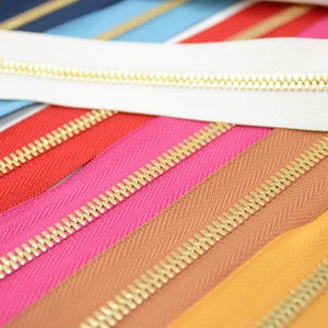 Puede incluir: Una colecci&oacute;n de cremalleras coloridas con dientes dorados. Las cremalleras son de varios colores, incluyendo blanco, rojo, rosa, marr&oacute;n y amarillo. La imagen es una toma de primer plano, destacando los detalles de los dientes y la tela de la cremallera.