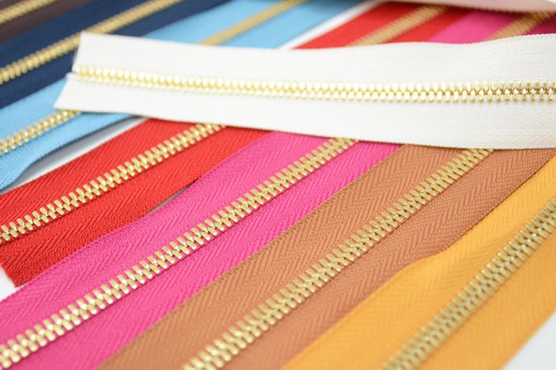 Puede incluir: Una colecci&oacute;n de cremalleras coloridas con dientes dorados. Las cremalleras son de varios colores, incluyendo blanco, rojo, rosa, marr&oacute;n y amarillo. La imagen es una toma de primer plano, destacando los detalles de los dientes y la tela de la cremallera.