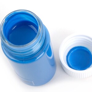May include: An overhead shot of a clear glass jar filled with bright blue paint, with the white lid beside it. The jar is partially transparent, revealing the blue paint inside. The lid is white with a blue interior.