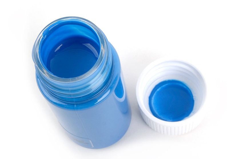 May include: An overhead shot of a clear glass jar filled with bright blue paint, with the white lid beside it. The jar is partially transparent, revealing the blue paint inside. The lid is white with a blue interior.