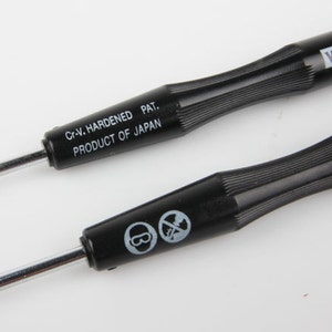 May include: Close-up of two black screwdrivers with silver tips. The handles are black with a ribbed design. Text on the handles reads "Cr-V. HARDENED PAT. PRODUCT OF JAPAN" and "VESSEL".