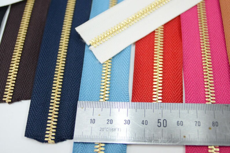 Puede incluir: Una colecci&oacute;n de cremalleras coloridas con dientes dorados. Las cremalleras son de varios colores, incluyendo marr&oacute;n, azul marino, azul claro, rojo, rosa y blanco. Una regla muestra una medida de 20 cm (68&deg;F).