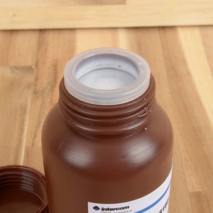 May include: A brown plastic bottle with a white interior and a clear plastic lid. The bottle is open, revealing the contents. The lid is off to the side. The bottle is on a wooden surface.