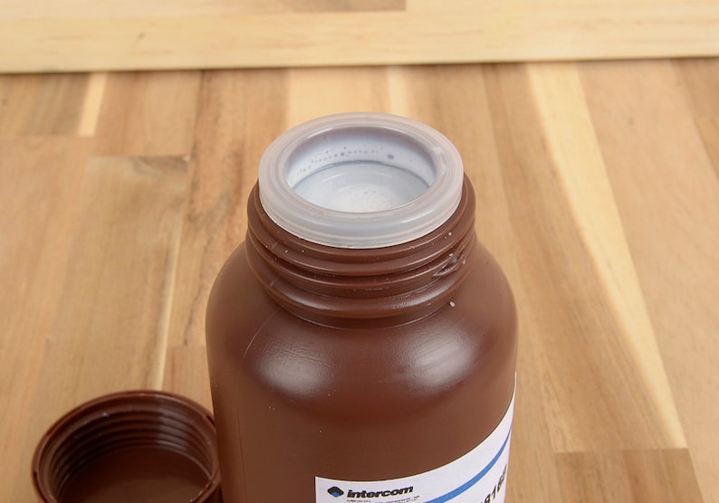 May include: A brown plastic bottle with a white interior and a clear plastic lid. The bottle is open, revealing the contents. The lid is off to the side. The bottle is on a wooden surface.