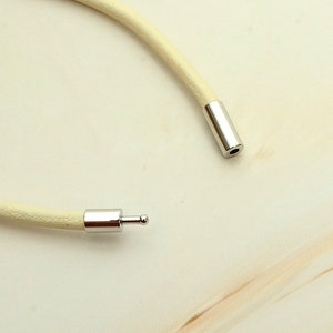 May include: A close-up of a cream-colored necklace with a silver clasp. The necklace is made of a soft, round material. The silver clasp is cylindrical with a small ball on one end. The background is a light cream color.
