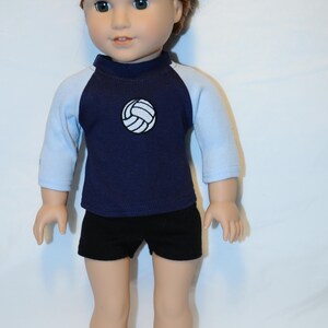 Puede incluir: Una mu&ntilde;eca con cabello casta&ntilde;o rojizo recogido en un mo&ntilde;o, que viste una camiseta azul marino con mangas ragl&aacute;n azul claro y un gr&aacute;fico de voleibol blanco en el pecho, junto con pantalones cortos negros lisos. La mu&ntilde;eca tiene tez clara y ojos verdes.