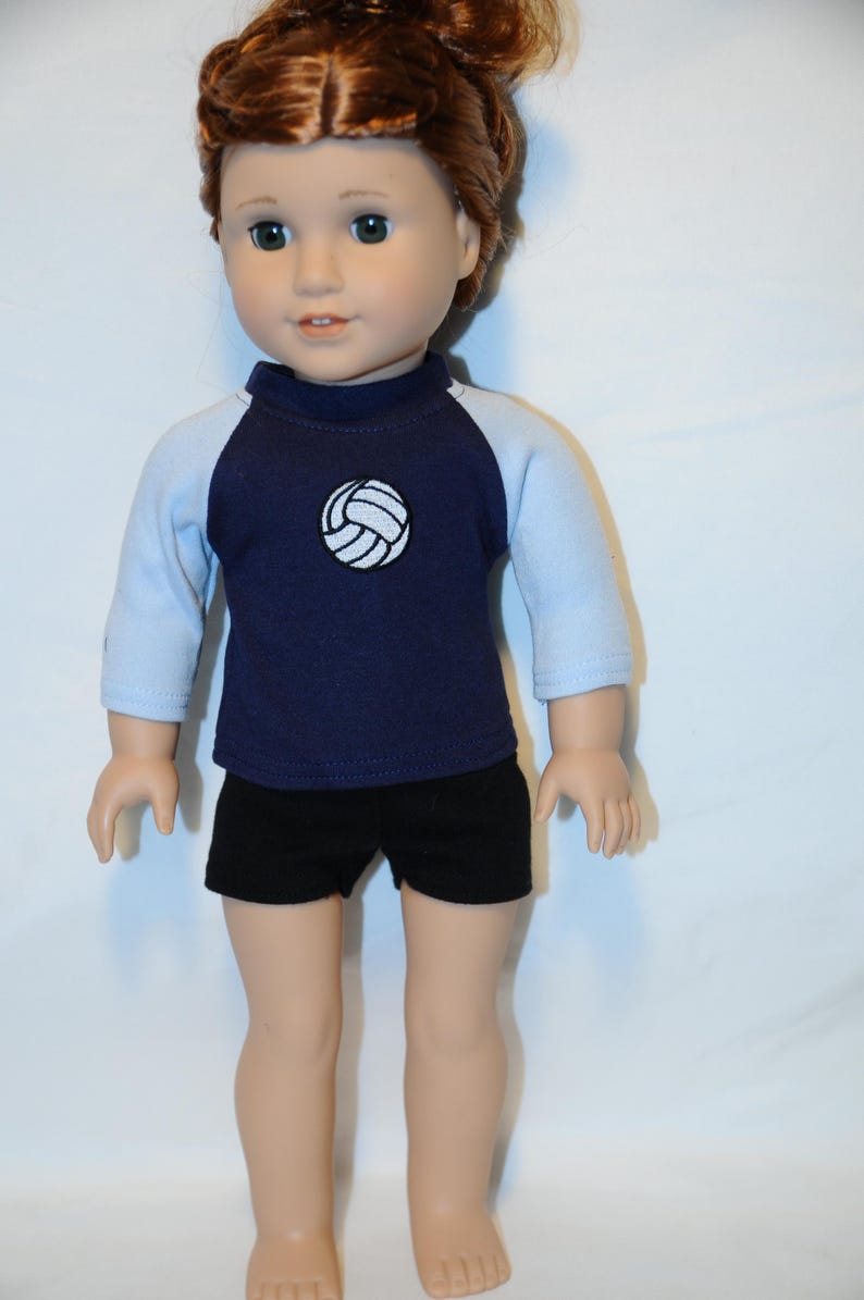 Puede incluir: Una mu&ntilde;eca con cabello casta&ntilde;o rojizo recogido en un mo&ntilde;o, que viste una camiseta azul marino con mangas ragl&aacute;n azul claro y un gr&aacute;fico de voleibol blanco en el pecho, junto con pantalones cortos negros lisos. La mu&ntilde;eca tiene tez clara y ojos verdes.
