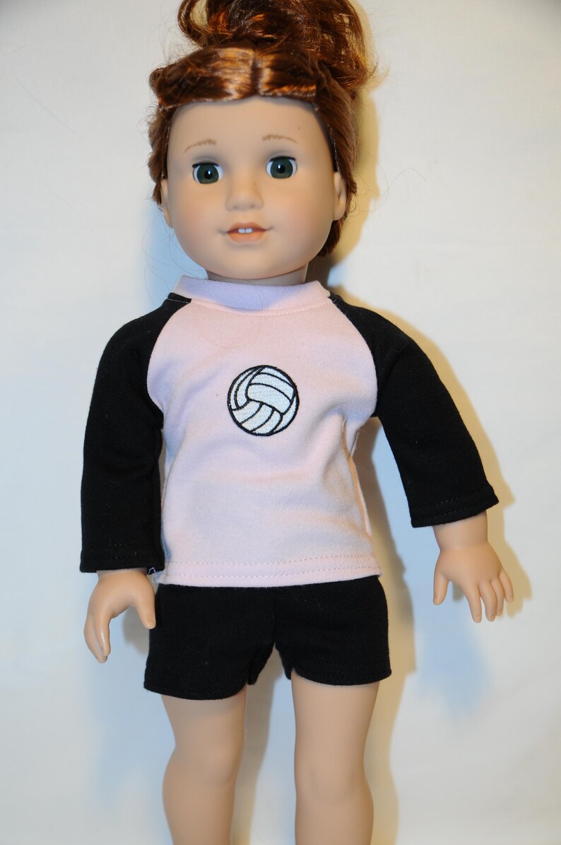 Puede incluir: Una mu&ntilde;eca con cabello casta&ntilde;o rojizo recogido en un mo&ntilde;o, que viste una camiseta de manga larga rosa claro con mangas ragl&aacute;n negras y un gr&aacute;fico de voleibol negro en el pecho, junto con pantalones cortos negros lisos. Tiene ojos verdes.