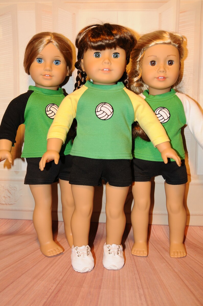 Puede incluir: Tres mu&ntilde;ecas de cabello casta&ntilde;o, cada una con una camiseta verde con un gr&aacute;fico de voleibol en el pecho y pantalones cortos negros. La mu&ntilde;eca de la izquierda tiene mangas negras, la del centro mangas amarillas y la de la derecha mangas blancas. La mu&ntilde;eca central lleva zapatos blancos.