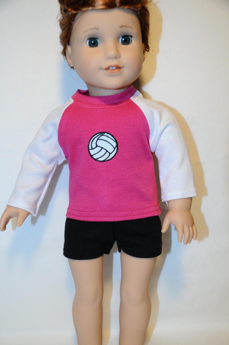 Puede incluir: Una mu&ntilde;eca de cabello casta&ntilde;o rojizo viste una camiseta de manga larga con torso rosa y mangas blancas, con un gr&aacute;fico de voleibol blanco centrado y contornos negros, combinado con pantalones cortos negros lisos hasta el muslo.