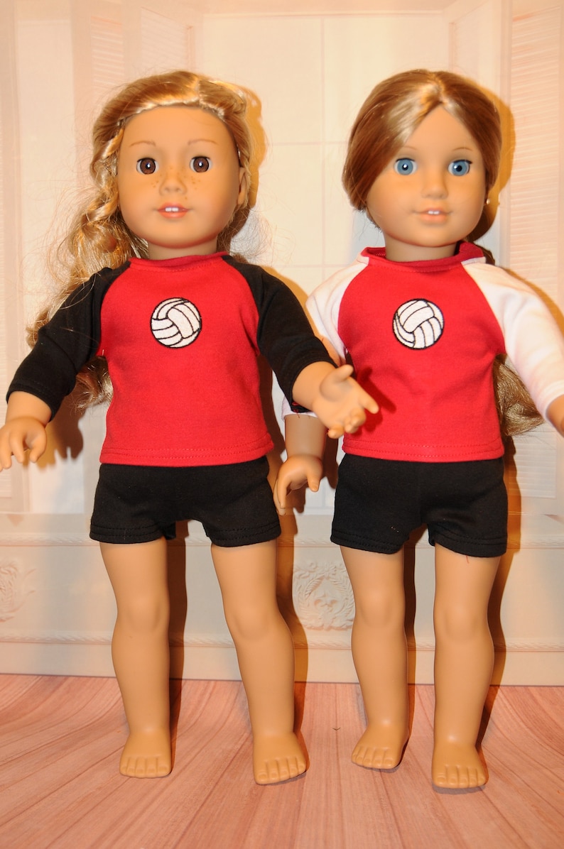 Puede incluir: Dos mu&ntilde;ecas de piel clara, una con cabello rubio ondulado y otra con cabello casta&ntilde;o claro liso. Ambas visten camisetas rojas con un gr&aacute;fico de voleibol blanco en el pecho, pantalones cortos negros y mangas ragl&aacute;n en color negro o blanco.