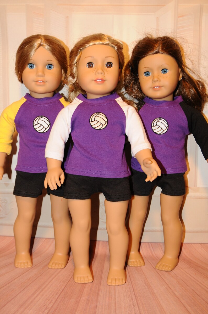 Puede incluir: Tres mu&ntilde;ecas de piel clara y ojos azules o marrones, cada una con una camiseta morada con un gr&aacute;fico de voleibol blanco en el pecho y pantalones cortos negros. Las camisetas tienen mangas tres cuartos de color amarillo, blanco y negro.