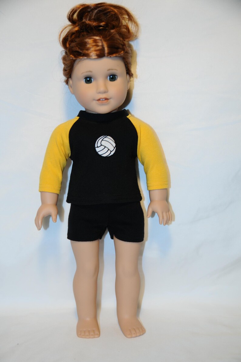 Puede incluir: Una mu&ntilde;eca con cabello rojizo recogido en un mo&ntilde;o, que viste una camiseta negra con mangas amarillas de tres cuartos y un gr&aacute;fico de voleibol blanco en el pecho, junto con pantalones cortos negros. La mu&ntilde;eca tiene piel clara y ojos oscuros.